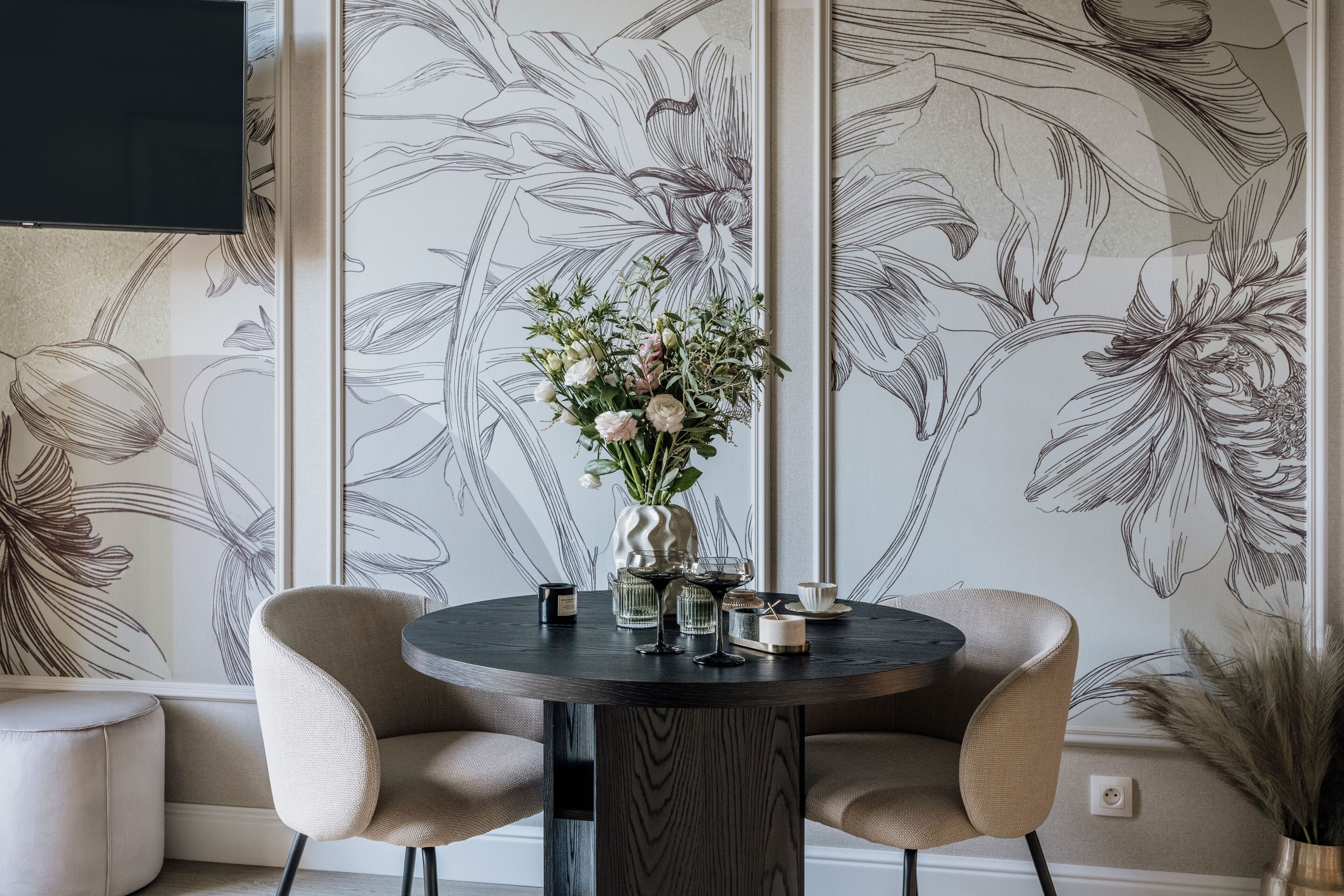 Zona pranzo con tavolo rotondo, due sedie, un vaso con fiori e parete decorata con motivo floreale.
