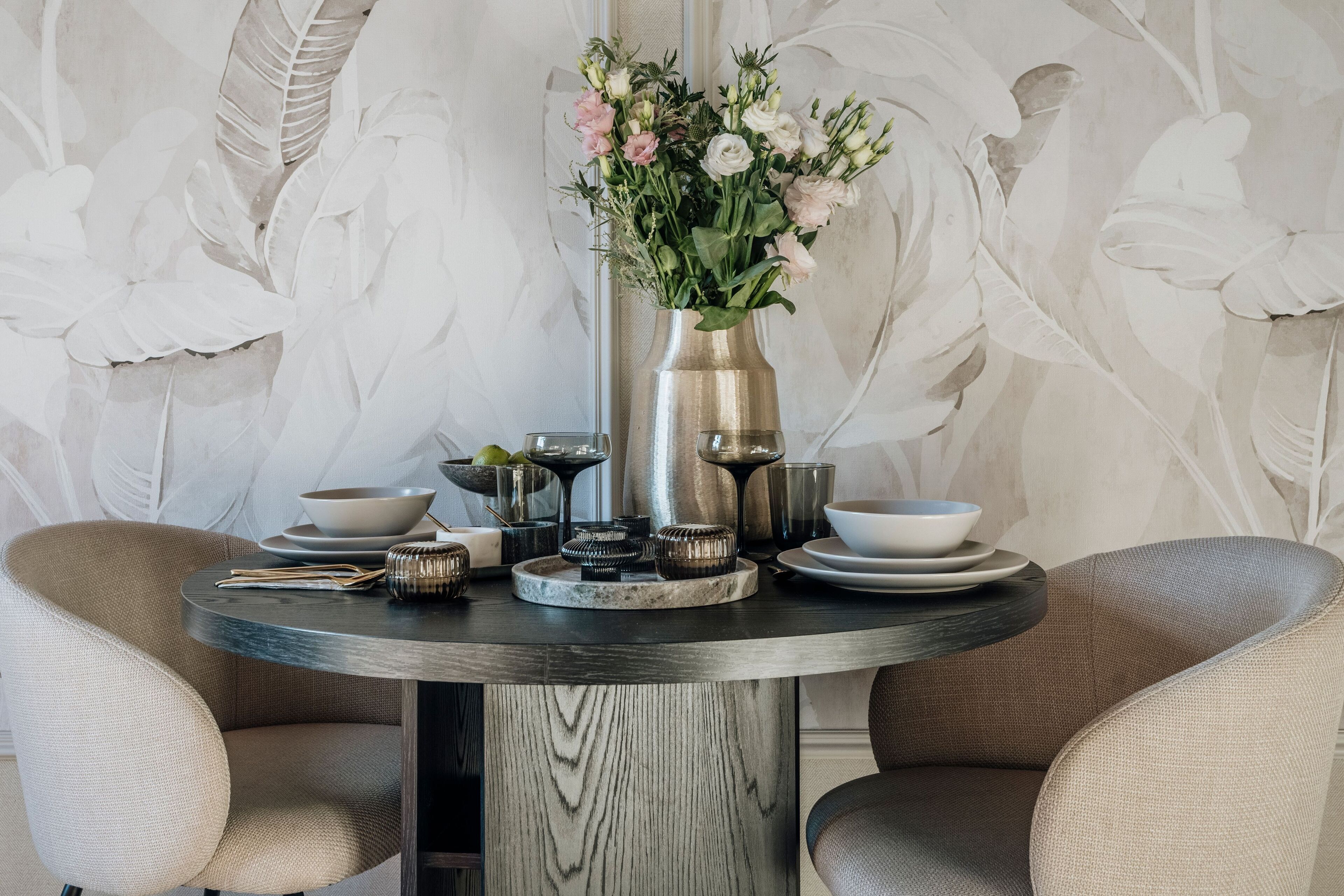 A round dining table with a vase of flowers, surrounded by chairs and various tableware.