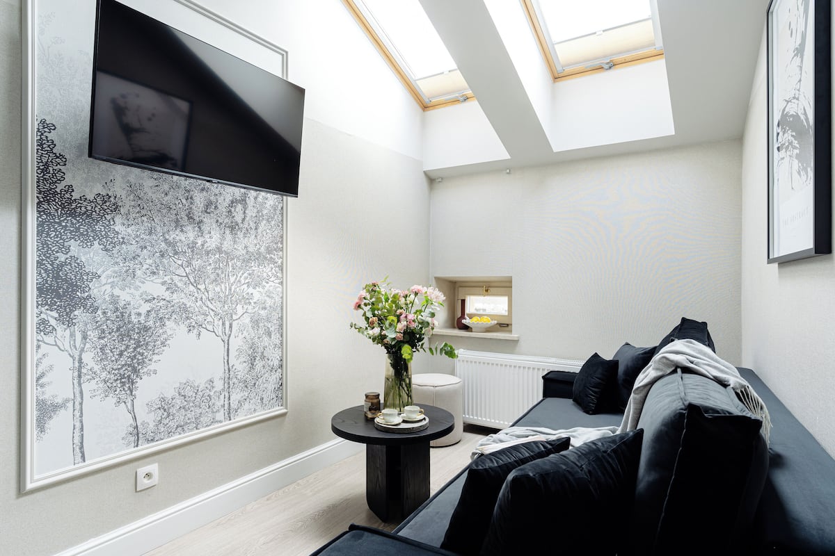 Un salon moderne avec un plafond haut, un téléviseur à écran plat fixé au mur, une table basse ronde ornée d’un vase de fleurs, un canapé noir avec des coussins et une fenêtre donnant sur des arbres.