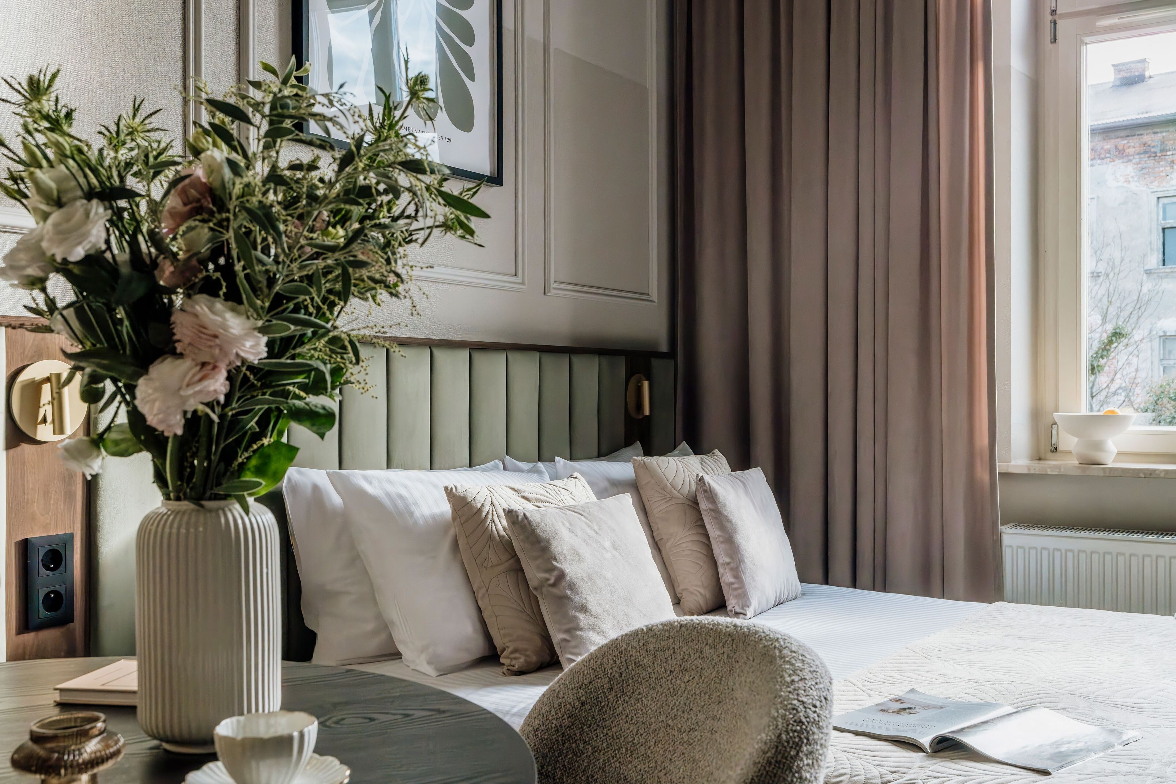 Habitación de hotel impecable con una cama, un florero, una mesita de noche con un libro y una ventana con cortinas.