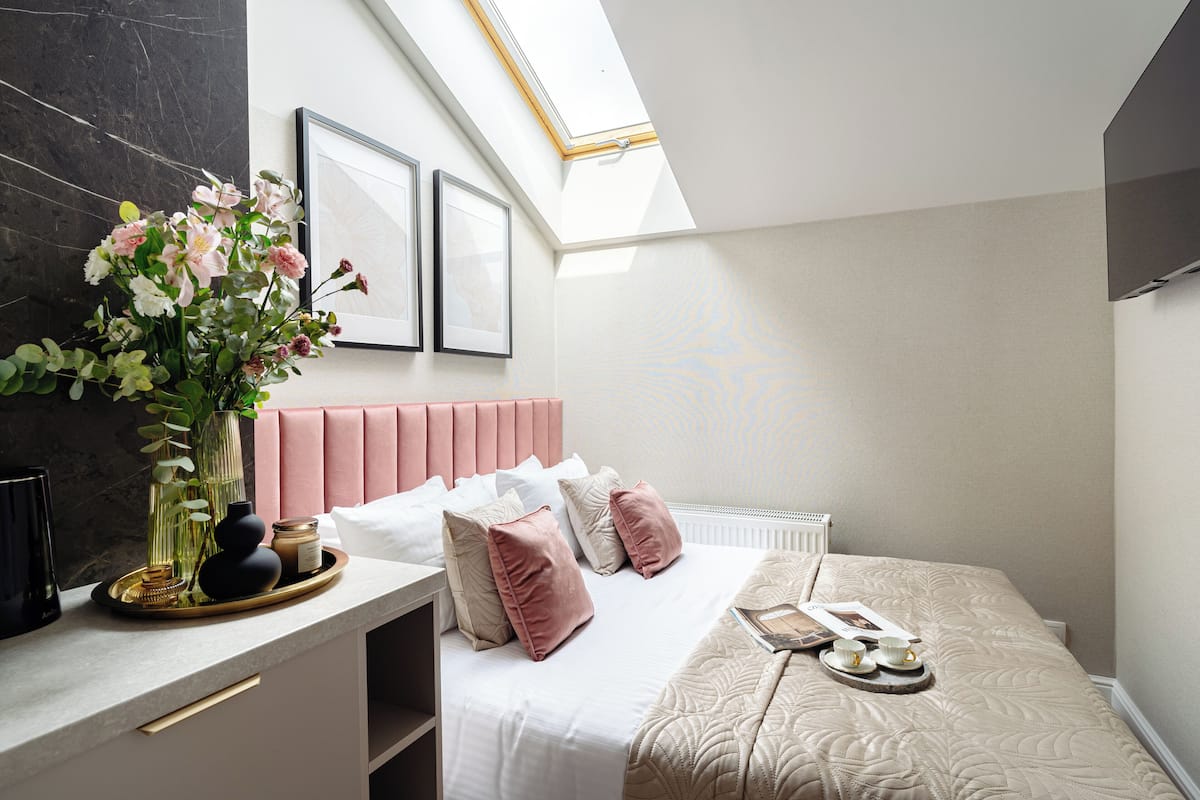 Une chambre moderne avec une lucarne, une table de chevet ornée d’un vase de fleurs, un lit avec une literie rose et blanche, et un tableau encadré accroché au mur.