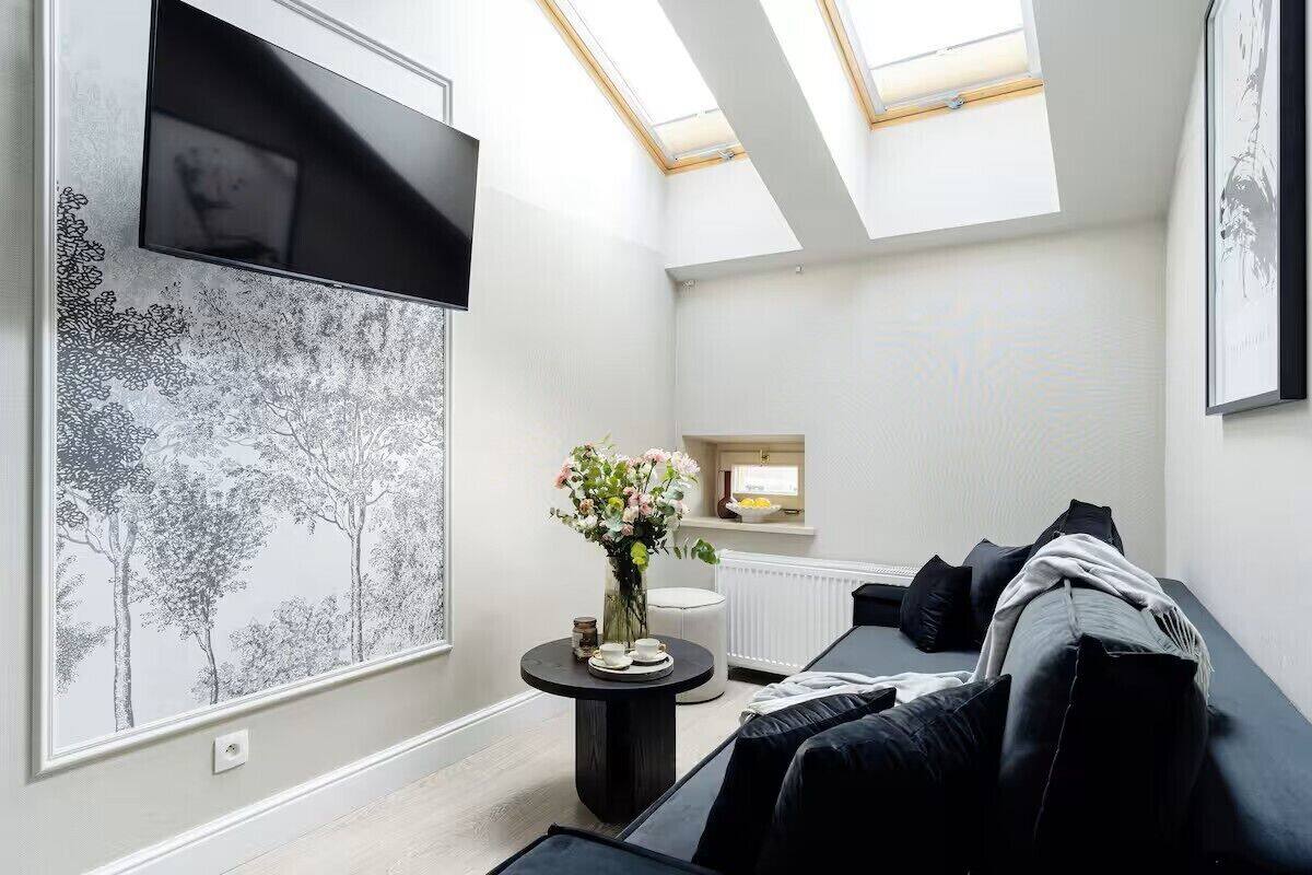 Un salon moderne avec un canapé noir, une table ronde ornée d’un vase de fleurs, un téléviseur à écran plat fixé au mur et une lucarne au plafond.
