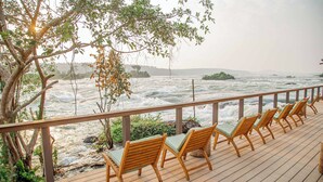A wooden deck with chairs overlooking a turbulent river, surrounded by greenery and a distant island.