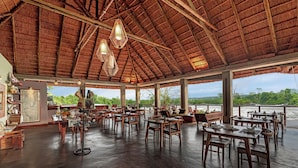 A spacious indoor dining area with wooden tables and chairs, a thatched roof, and large windows overlooking a scenic landscape.