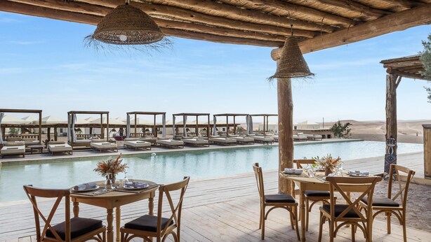 Area piscina con sedie e tavoli in legno, una lampada a sospensione e vista sulla piscina e sul paesaggio desertico.