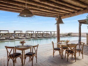 A poolside area with wooden deck chairs and tables, a hanging light fixture, and a view of a pool and desert landscape. - Les Terrasses d'Agafay (Sidi Badhaj)