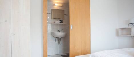 A bedroom with a wooden door leading to a bathroom. The room has a bed, a small shelf, and a wall-mounted light fixture.