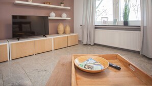 Ein modernes Wohnzimmer mit Fernseher, Holzschränken, einer Ablage mit Zeitschriften und einem Fenster mit Vorhängen.