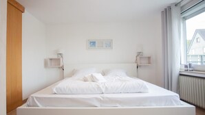 Ein schlicht eingerichtetes Schlafzimmer mit einem großen Bett, weißer Bettwäsche, zwei Nachttischen und einem Fenster mit Blick auf Gebäude.