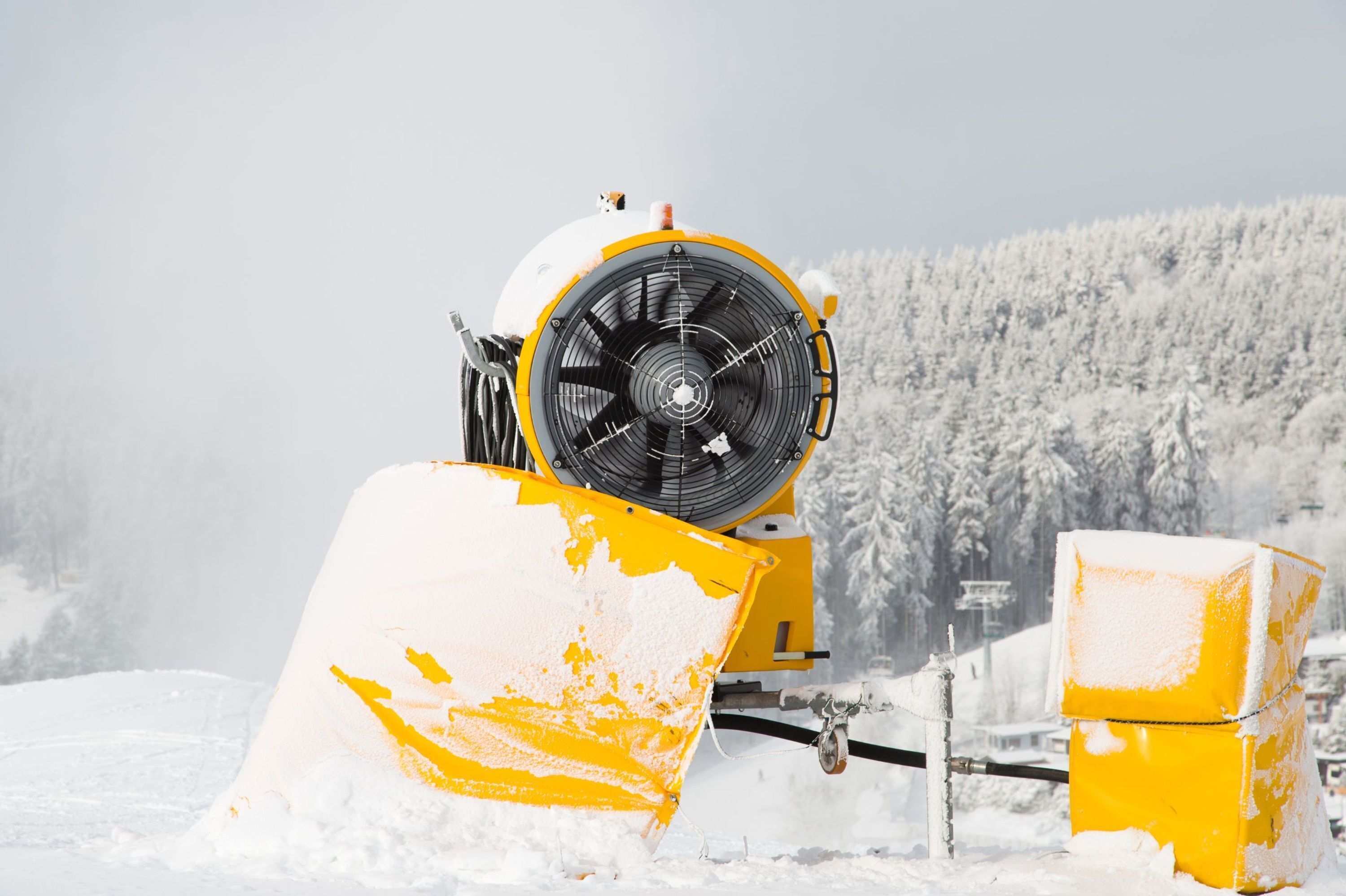 A snow-covered yellow machine with a large fan, set against a snowy forest backdrop.