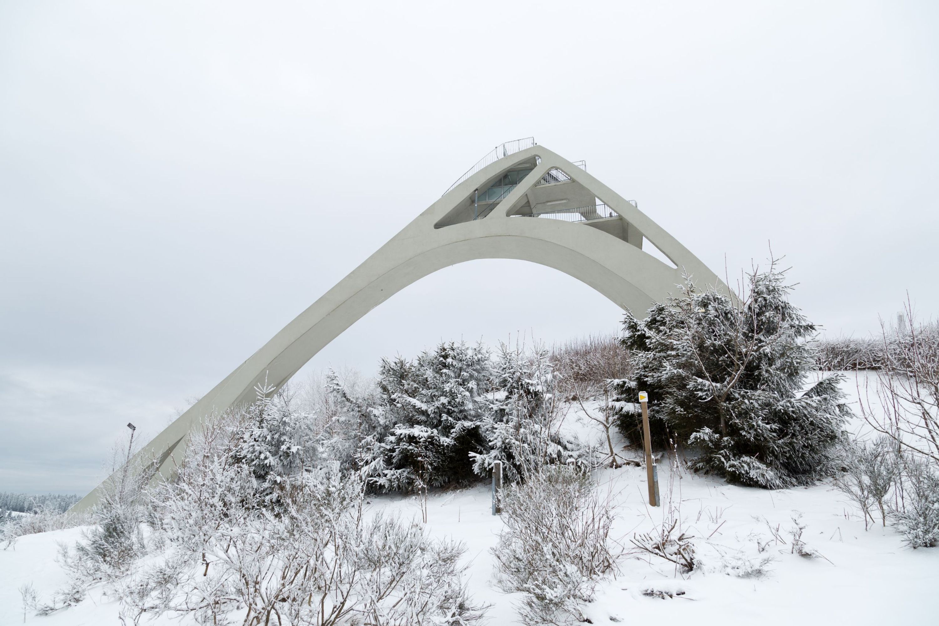 A large, white, arched bridge structure amidst a snowy landscape with sparse vegetation.
