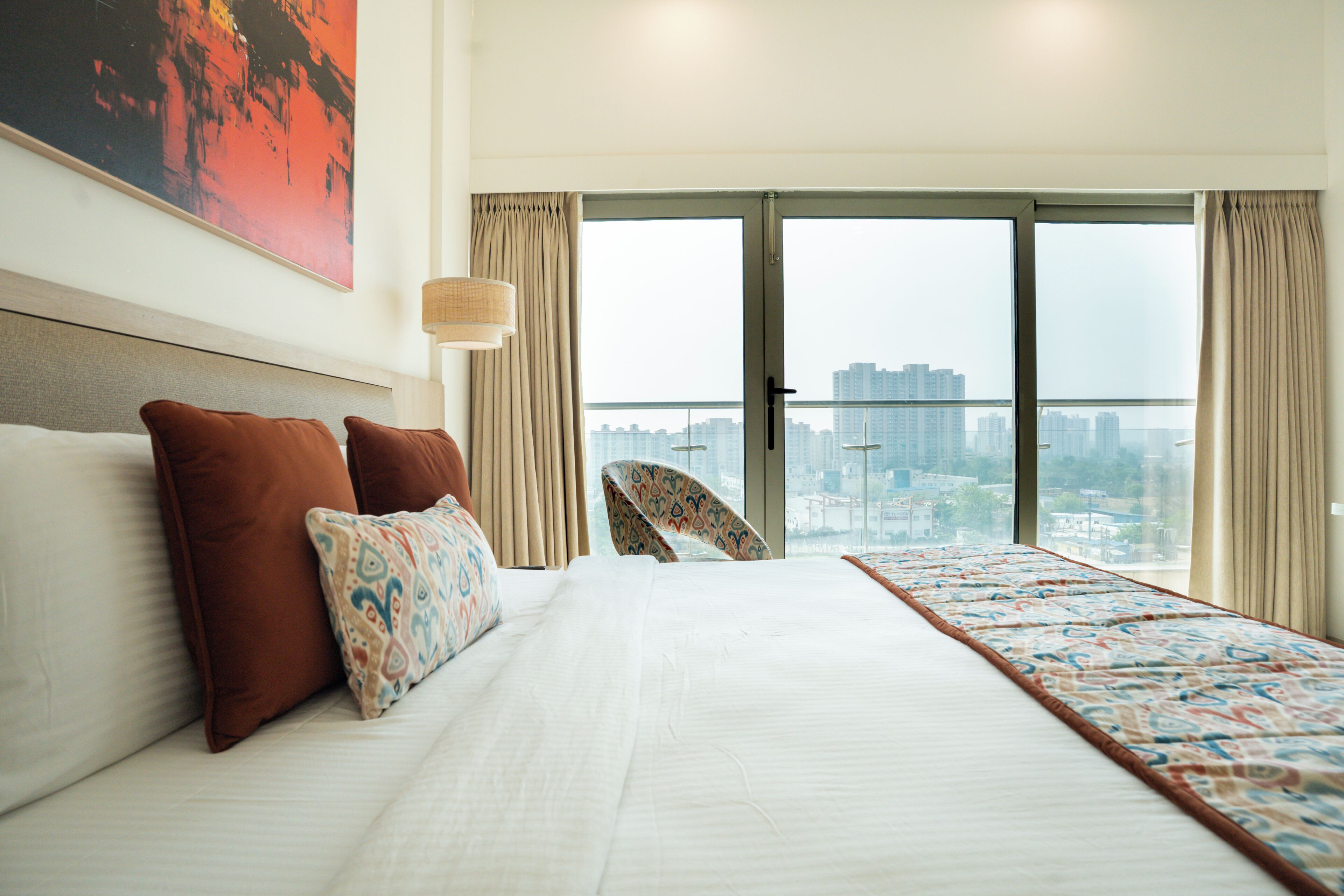 A hotel room with a large bed, two pillows, a patterned throw, a city view through the window, and a chair near the window.