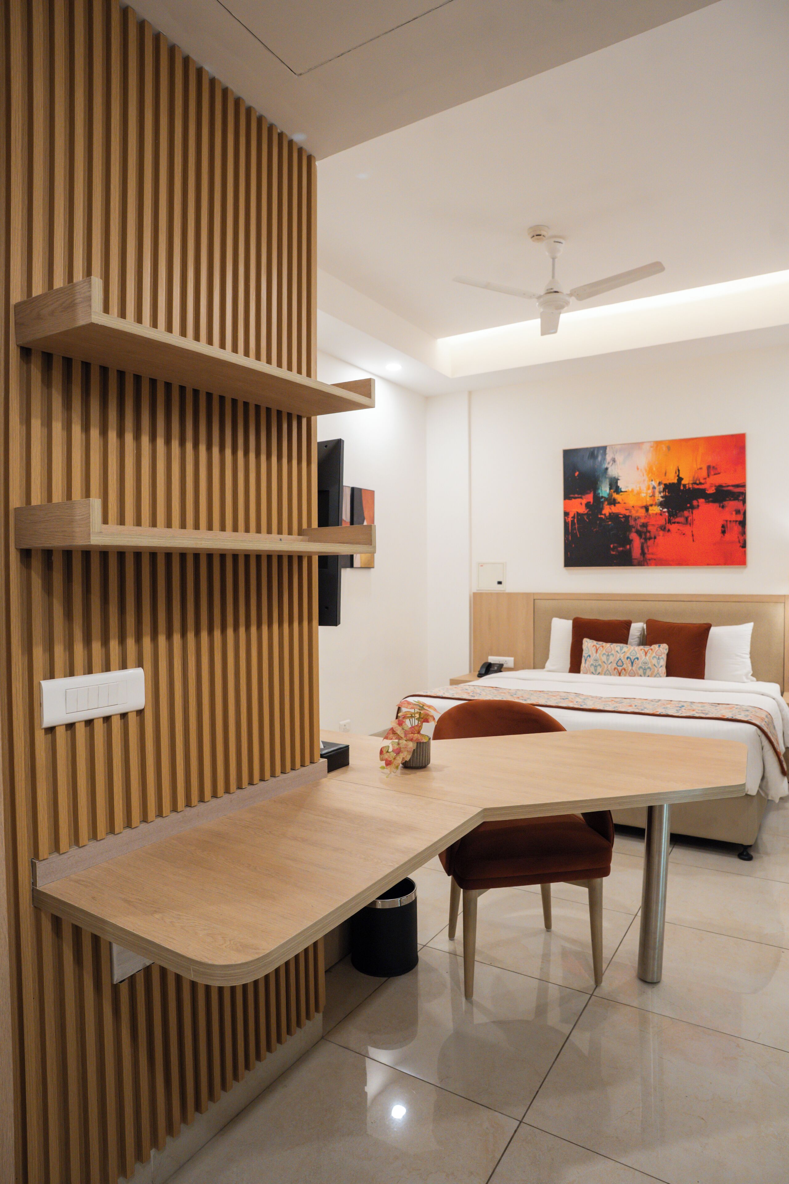 A modern hotel room with a wooden slatted wall, a desk with a chair, a bed with a headboard, a ceiling fan, and a painting on the wall.