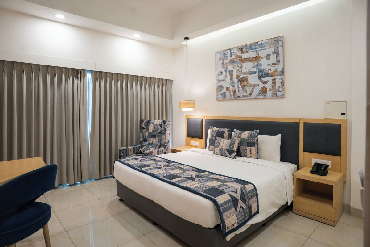 A hotel room with a large bed, a desk, a chair, a nightstand with a phone, and a painting on the wall.