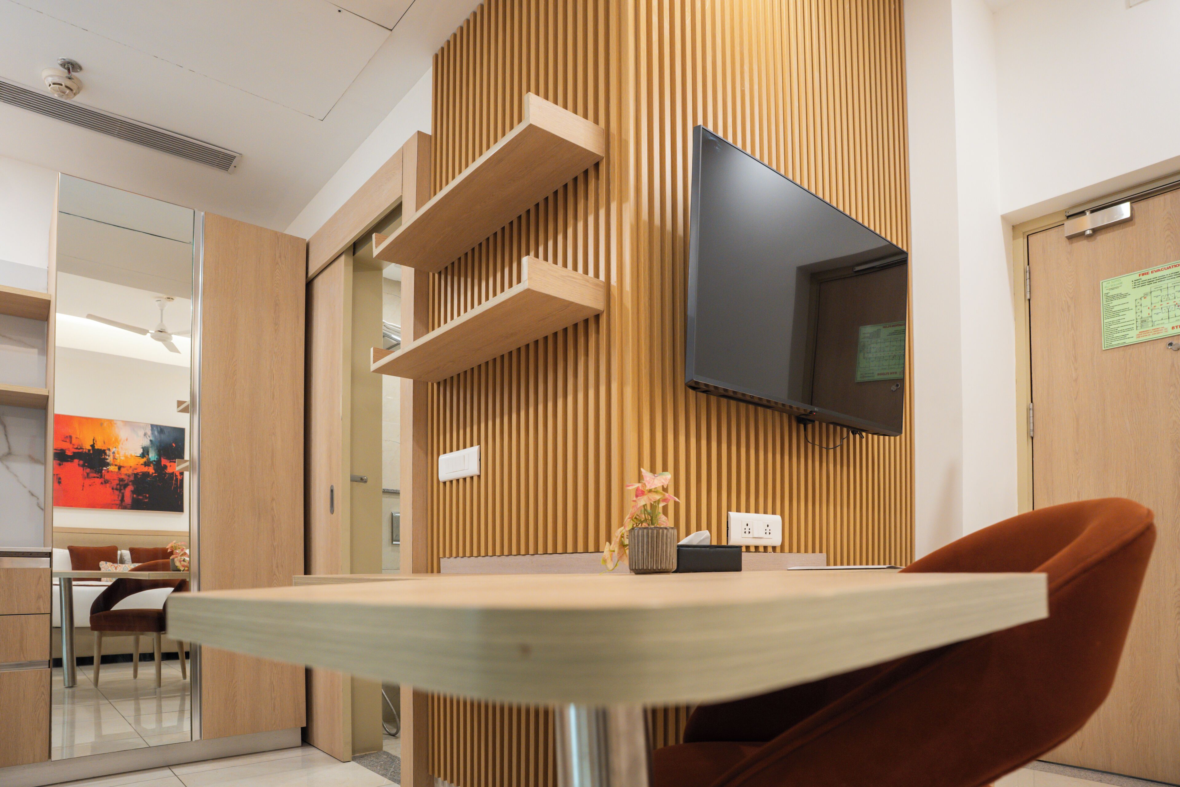 A modern interior with a wooden panel wall, a flat-screen TV mounted on the wall, a dining area with a table and chairs, and a reception desk.