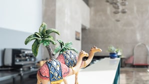 Kameli-aiheinen koriste-esine, jossa on satula ja palmu, on sijoitettu valkoiselle tasolle moderniin sisustustilaan. Tilaan kuuluu kiviseinä ja siitä roikkuu pallomaisia valaisimia.