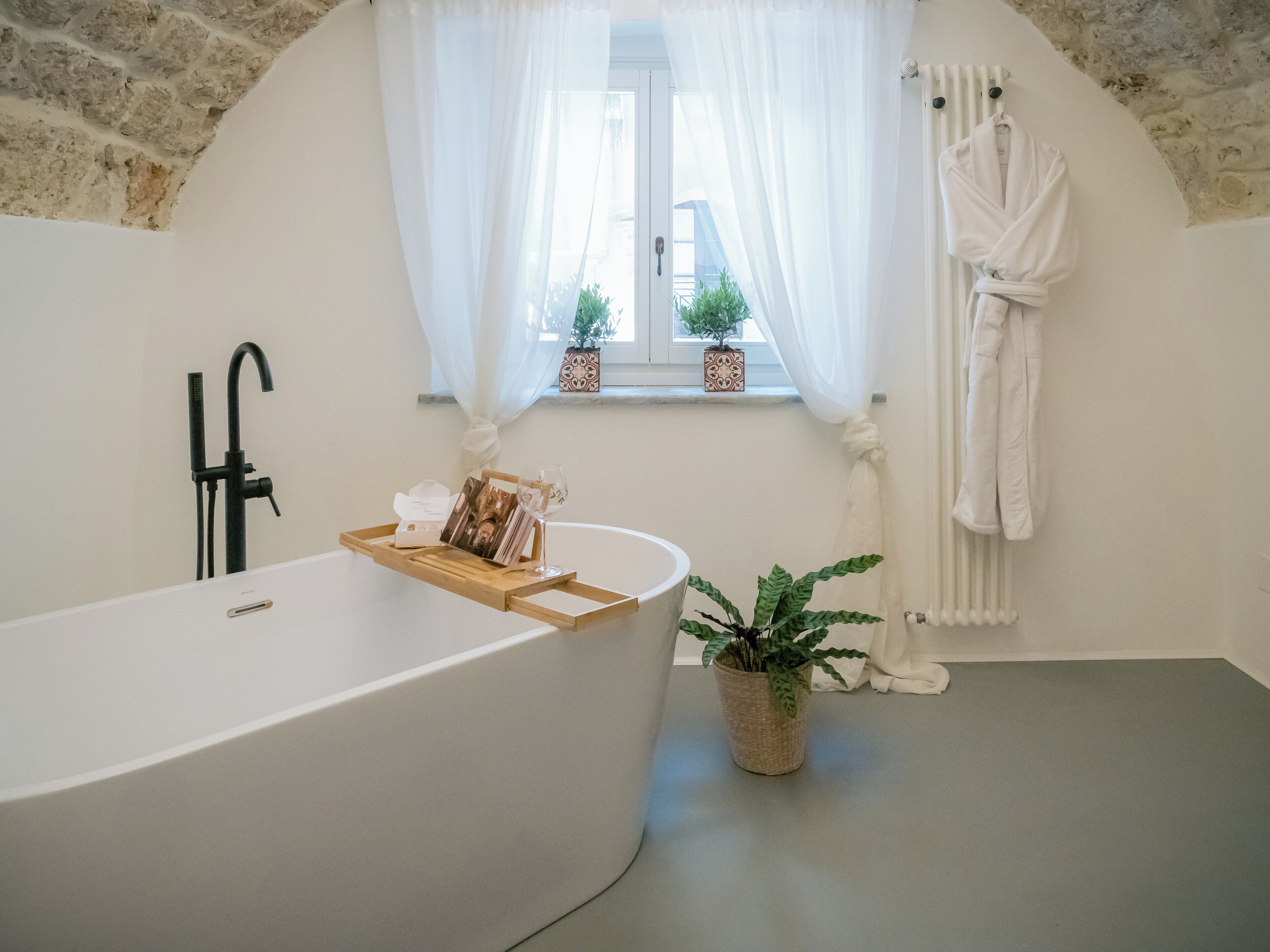 A modern bathroom with a freestanding bathtub, a window with sheer curtains, a potted plant, and a towel rack.