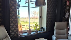 Ein Zimmer mit Sofa, Sessel, kleinem Tisch mit Fernbedienung, Fenster mit Blick ins Freie und einer Lampe.