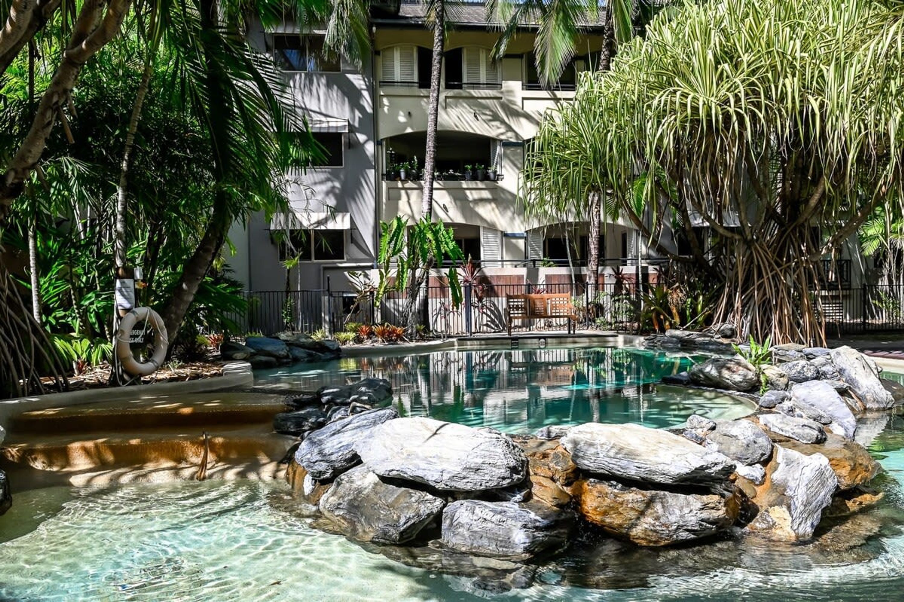 A hotel with a swimming pool surrounded by rocks and plants, a palm tree, and a building with balconies.