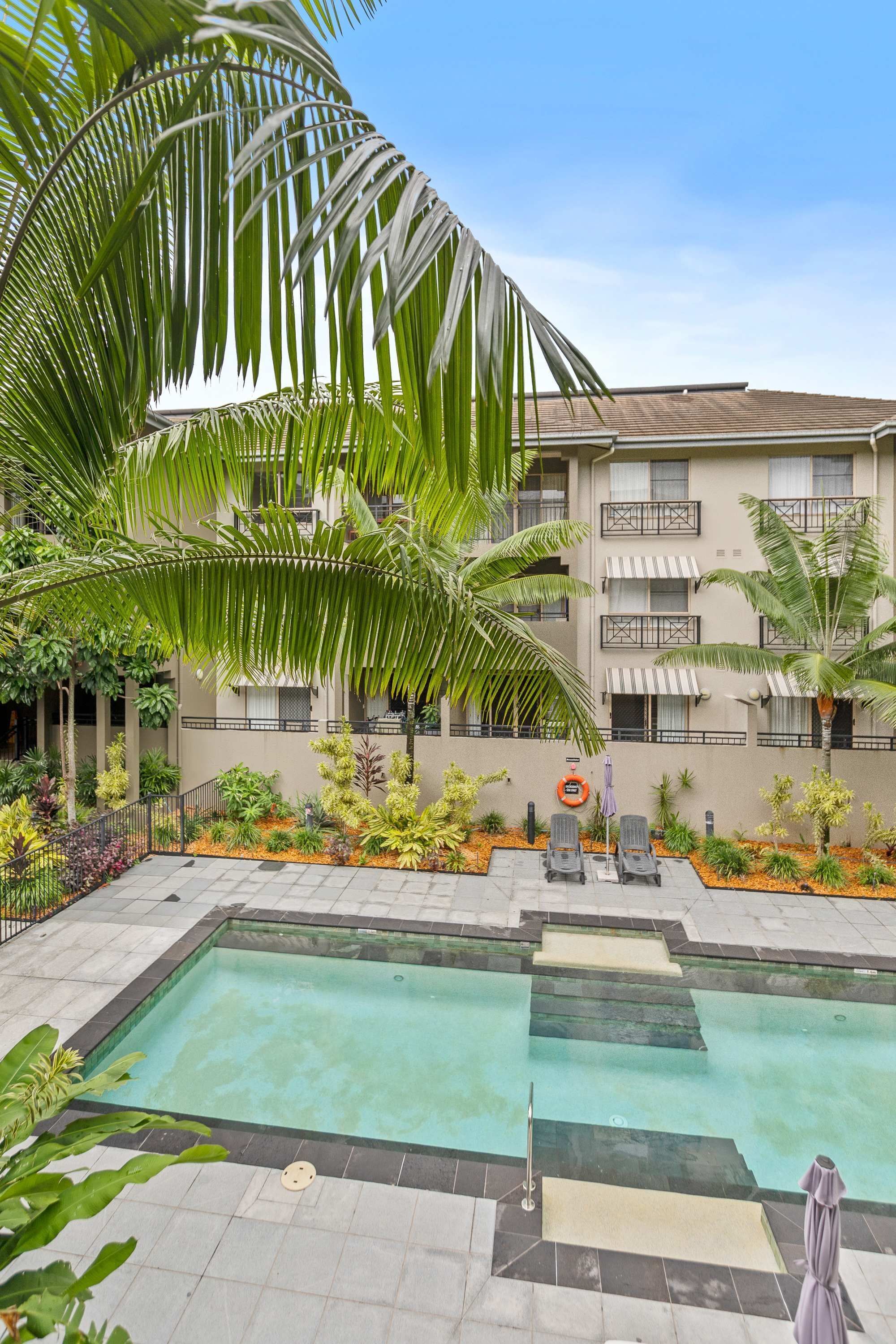 A swimming pool with lounge chairs, surrounded by palm trees and various plants, with a building in the background.