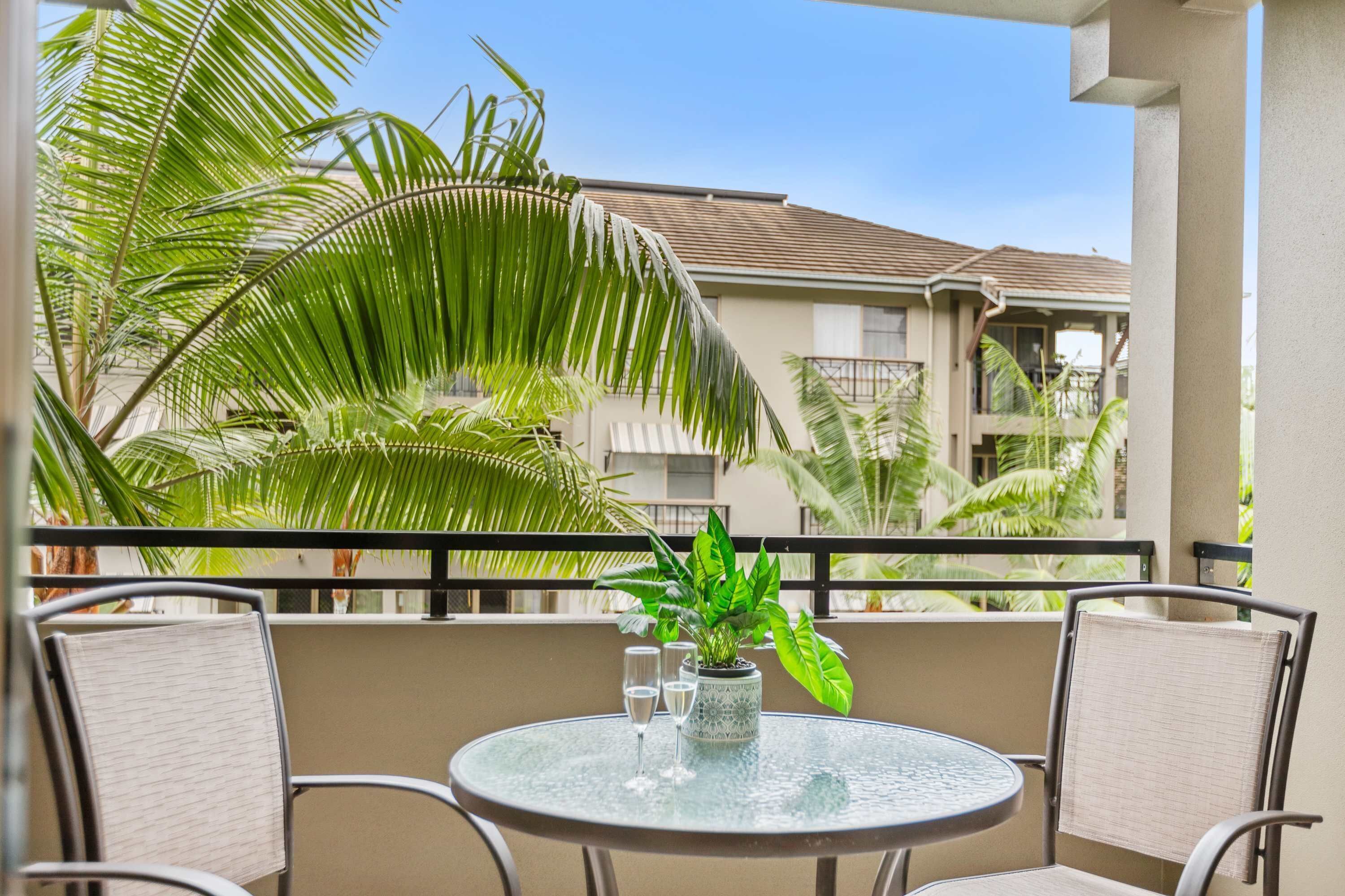 A round glass table with chairs, a potted plant, and two glasses on a balcony with a view of a building and palm trees.