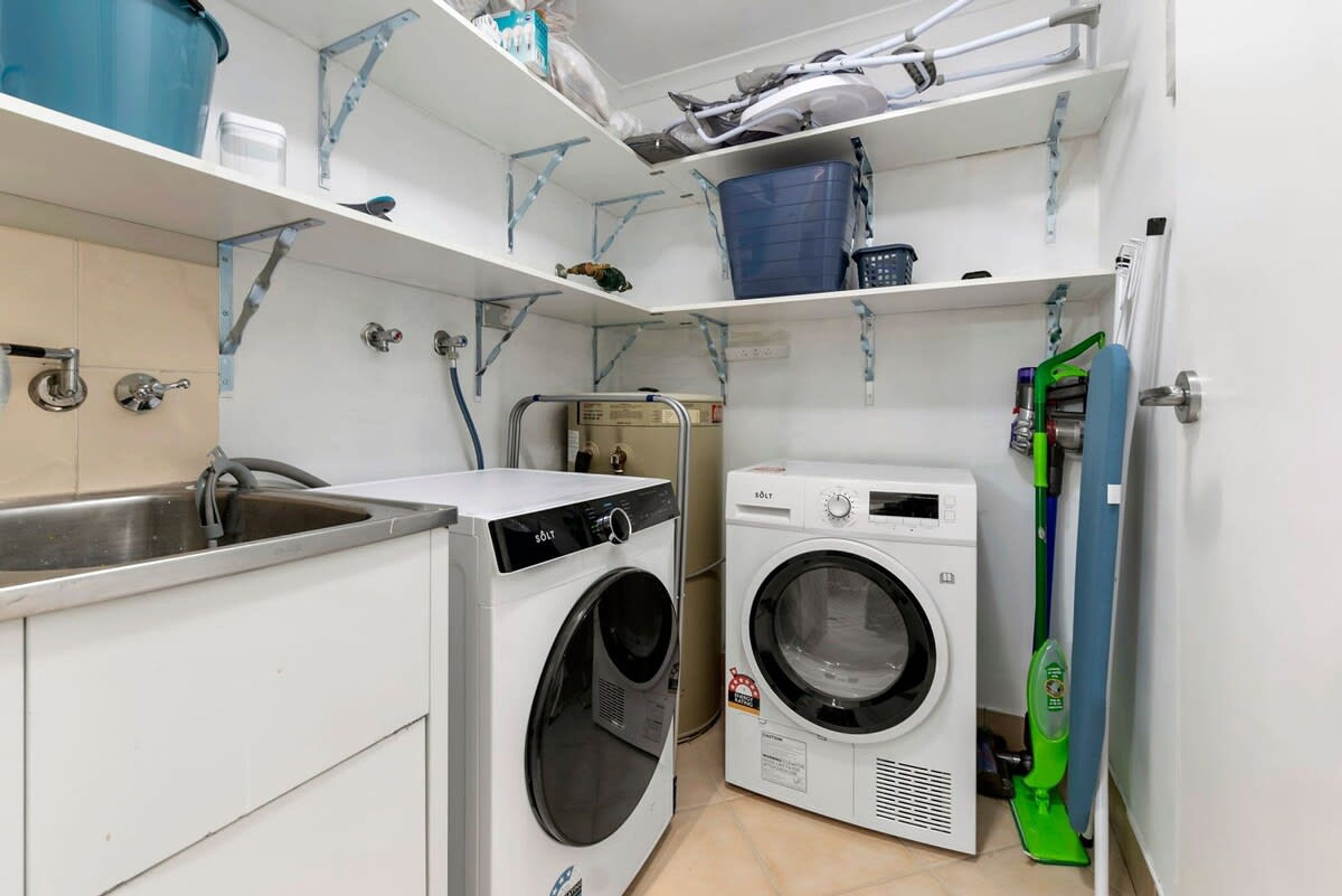 Pequeño cuarto de lavandería con dos lavadoras, un lavabo y estantes con diversos objetos.