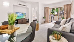 A modern living room with a glass dining table, grey sofa, and a TV on the wall. - Lux Apartment Cairns - 9 Pools BBQ Gym (Westcourt)