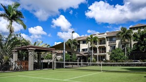 A tennis court with a net, surrounded by a fence, and adjacent to a building with balconies and palm trees.