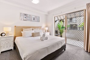 A neatly made bed with white linens and a folded towel, a bedside table with a lamp, and a large window with a view of the outdoors. - Lux Apartment Cairns - 9 Pools BBQ Gym (Westcourt)