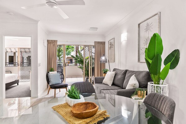A modern living room with a glass coffee table, grey sofa, and large green plants. - Lux Apartment Cairns - 9 Pools BBQ Gym (Westcourt)
