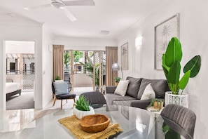 A modern living room with a glass coffee table, grey sofa, and large green plants.