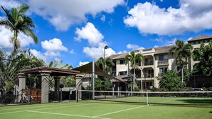 Una cancha de tenis con red, rodeada por una valla, palmeras y edificios al fondo.