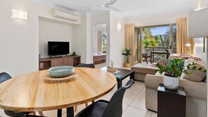 A modern living room with a wooden dining table, a sofa, and a television. - Poolside Paradise Cairns - 9 Pools BBQ Gym (Westcourt)