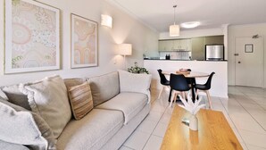 A modern living room with a sofa, a dining table, and a kitchen in the background. - Poolside Paradise Cairns - 9 Pools BBQ Gym (Westcourt)