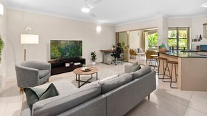 En moderne stue med sofa, lænestole, sofabord og fjernsyn. Der er et køkkenområde med bar og barstole.