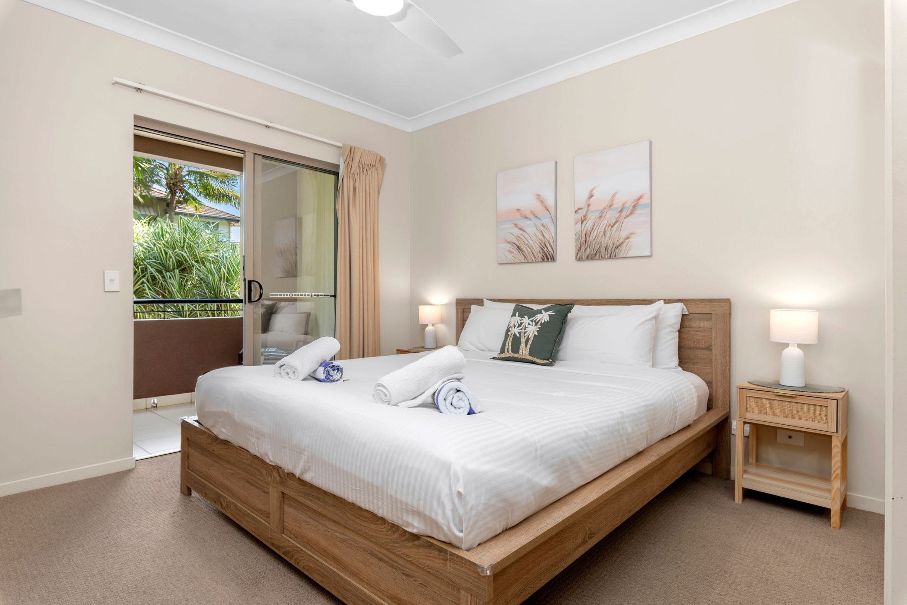 A bedroom with a wooden bed, two bedside tables with lamps, a sliding glass door to a balcony, and two wall-mounted artworks.