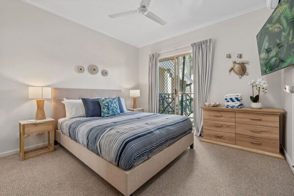 Une chambre à coucher comprenant un lit, des tables de chevet, une commode, un ventilateur de plafond et une vue par la fenêtre.