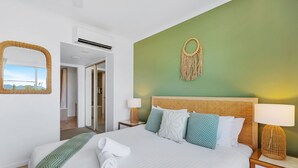 Un llit ben arropat amb llençols blancs i una manta verda, una làmpada de nit i una decoració de macramé penjada a la paret.