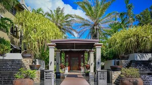 Une entrée abritée, dotée d’une porte en bois et flanquée de grands jardinières et de palmiers, mène à un bâtiment à la façade en pierre.