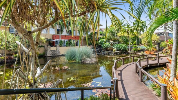 A tropical garden with a wooden bridge over a pond, surrounded by various plants and a building in the background. - The Nest Cairns - 4 Pools BBQ Resort (Cairns North)