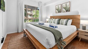 A bedroom with a large bed, a wooden headboard, a nightstand, a window with curtains, and two wall-mounted pictures. - The Nest Cairns - 4 Pools BBQ Resort (Cairns North)