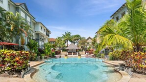 A pool area with a clear blue pool, surrounded by palm trees and a building with balconies. - The Nest Cairns - 4 Pools BBQ Resort (Cairns North)