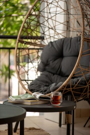 A wicker chair with a cushion, a table with a book and a glass of tea, and a view of greenery outside.