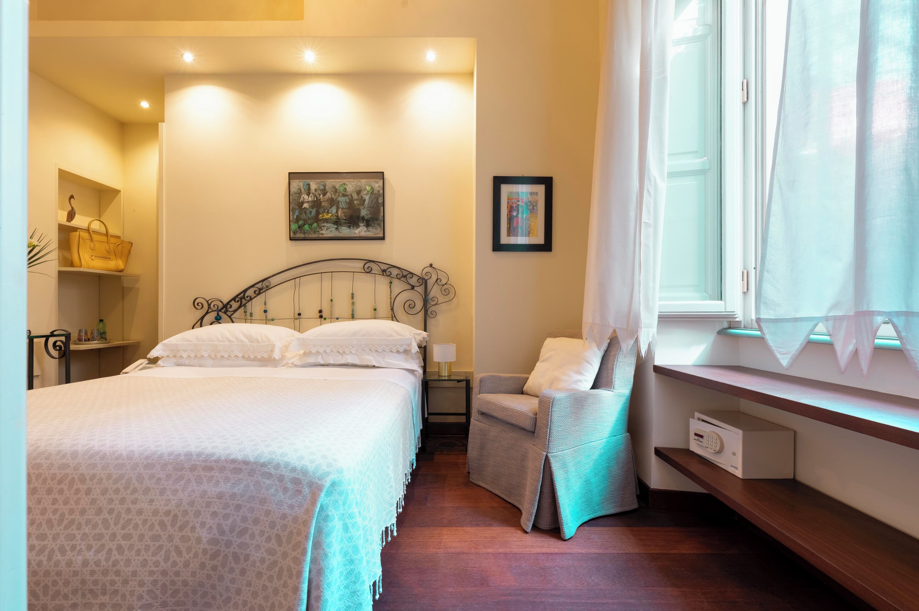 A bedroom with a bed, a chair, a nightstand, a lamp, a painting, and a window with curtains.