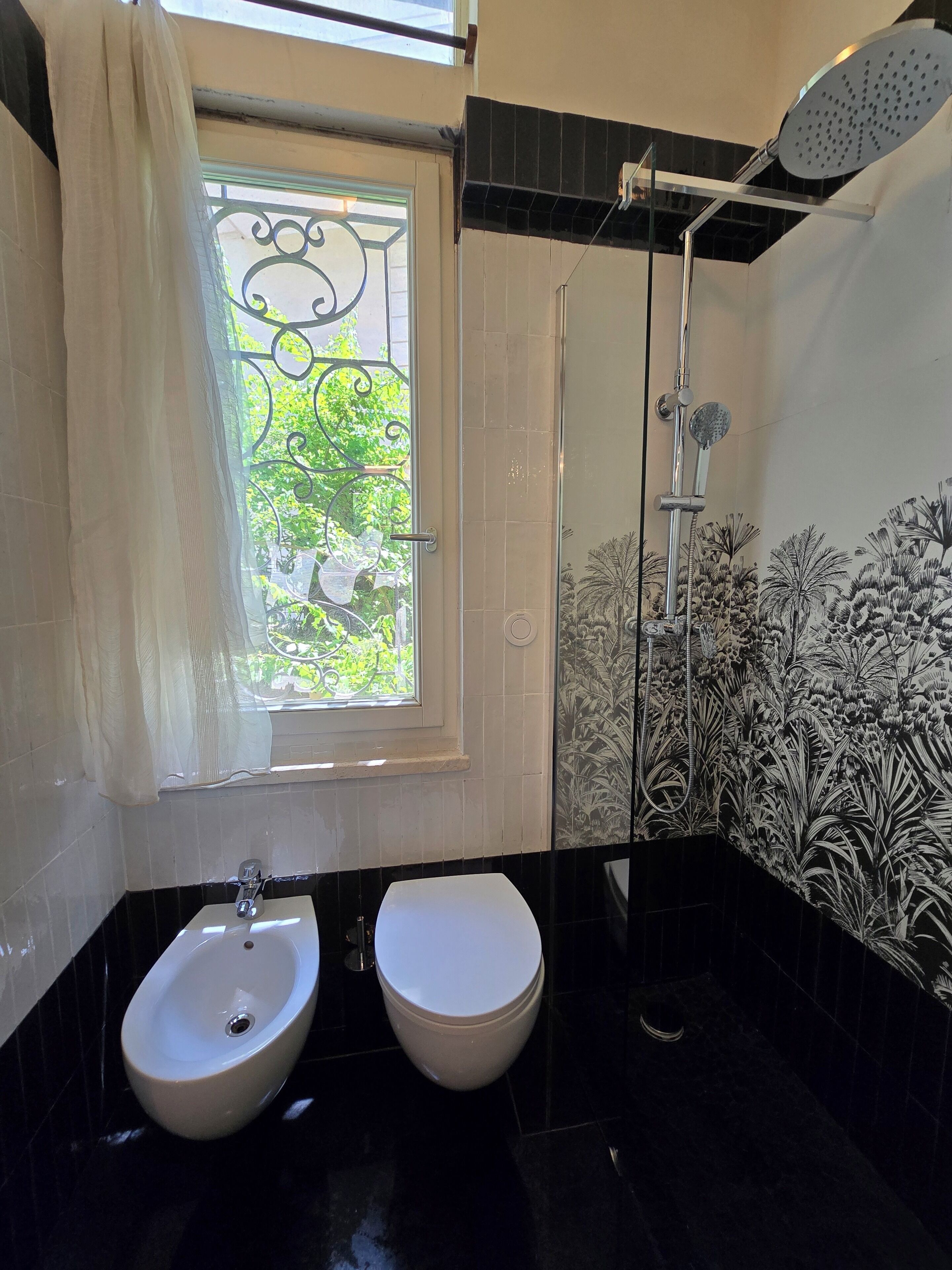 Une salle de bain équipée d’un bidet, de toilettes, d’une douche et d’une fenêtre donnant sur un espace verdoyant.