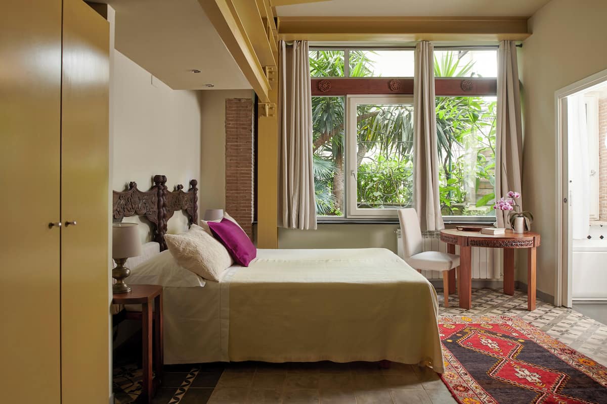 A bedroom with a bed, a nightstand, a chair, a small table, and a window with curtains.