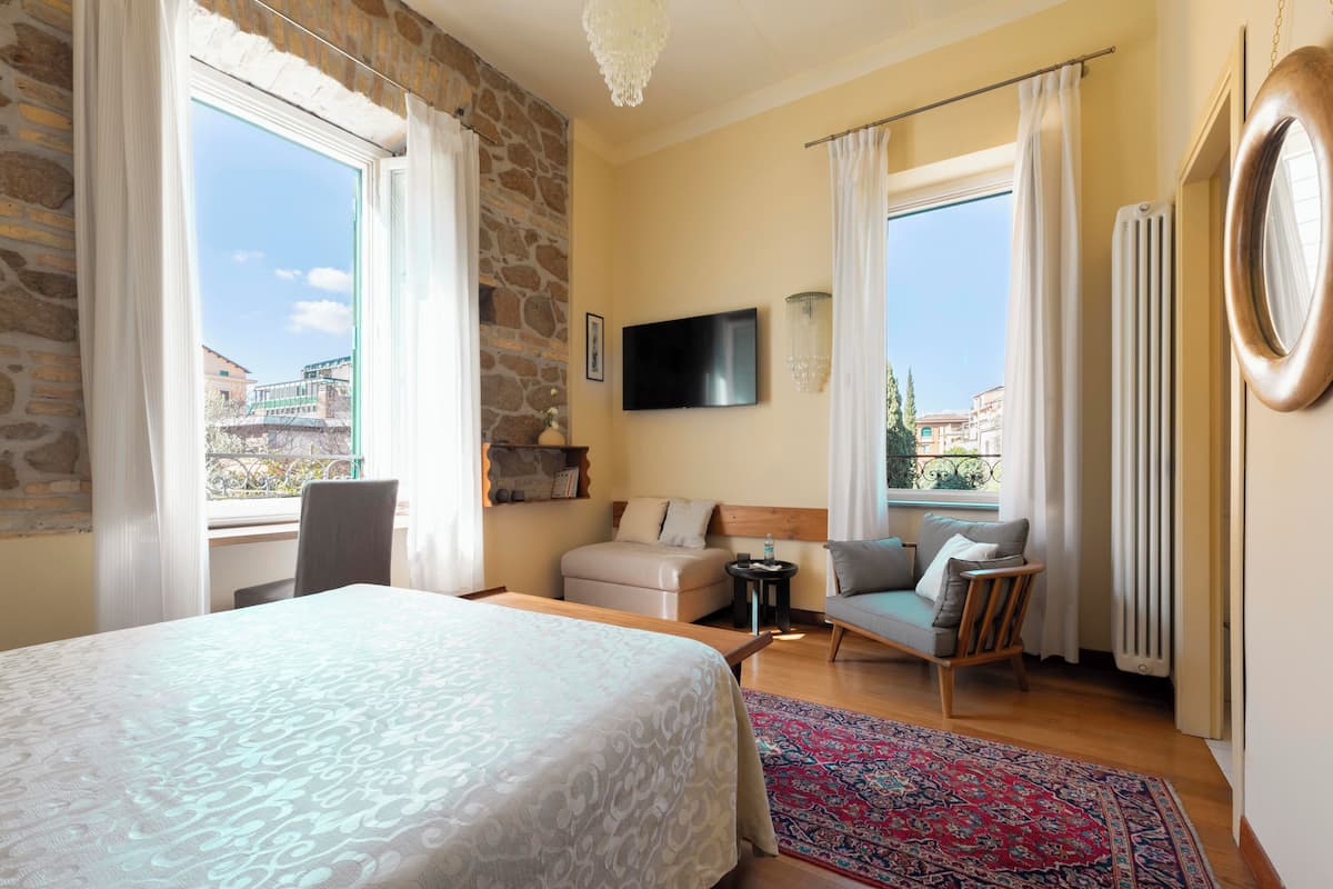 A hotel room with a bed, a sofa, a TV, and a view of buildings outside.
