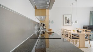 A modern kitchen with a central island, stainless steel appliances, and a dining area with wooden chairs and a table. - Gudhjem Søpark Lejl. 23 (Gudhjem)