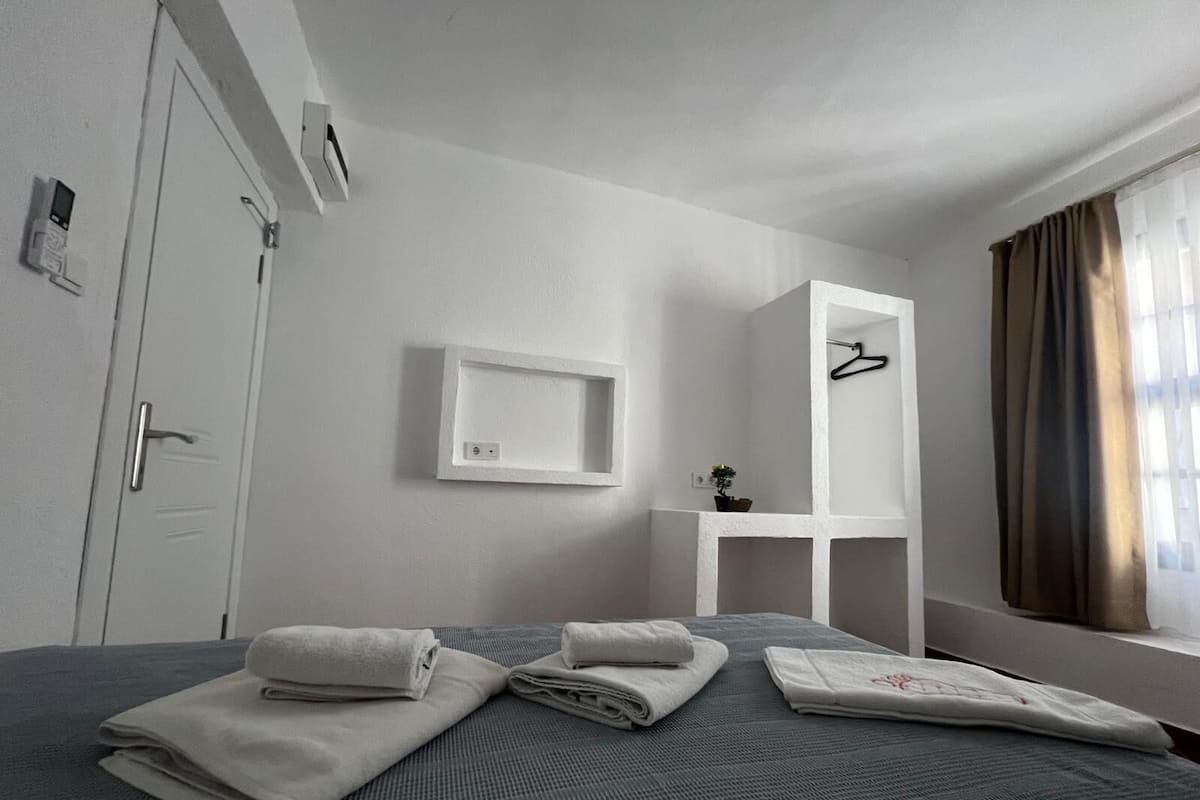 Quarto de hotel com cama, mesa de cabeceira, espelho, luminária pendente e janela com cortinas.