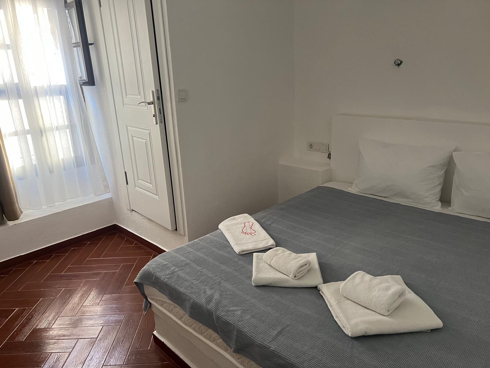A hotel room with a bed, a window with curtains, a door, and a wooden floor.